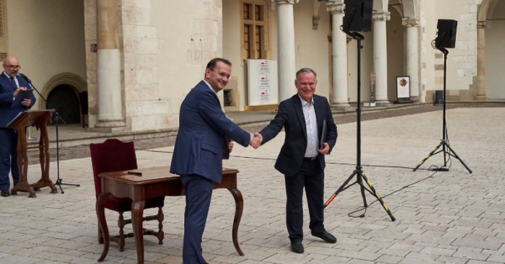 dziedziniec zamku, dwaj uśmiechnięci mężczyźni podają sobie ręce, za nimi stylowy mały stolik i krzesło, w tle arkady zamku